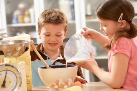 two children baking in a kitchen, one is stirring a bowl of mixture and the other is pouring a liquid from a jug