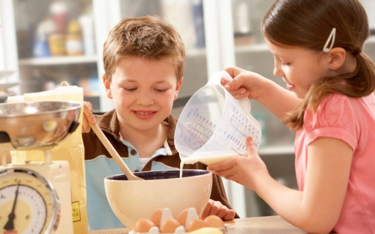 two children baking in a kitchen, one is stirring a bowl of mixture and the other is pouring a liquid from a jug