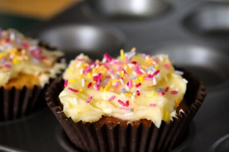 two iced fairy cakes covered in colourful sprinkles in a muffin tray