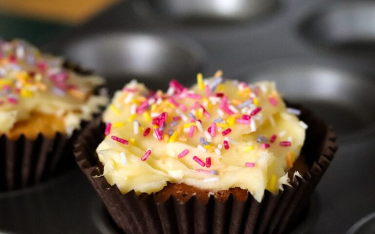 two iced fairy cakes covered in colourful sprinkles in a muffin tray